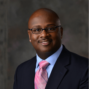 Deadwric Williams wears glasses and a suit as he poses against a professional background for a headshot