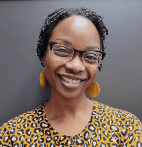Regina Baker smiles in glasses and yellow circular earrings in a headhsot