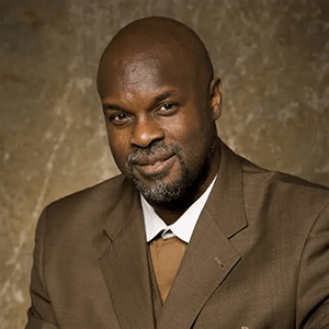 Tukufu Zuberi wears a brown suit with brown tie as a smiles against a professional backgroun for his headshot